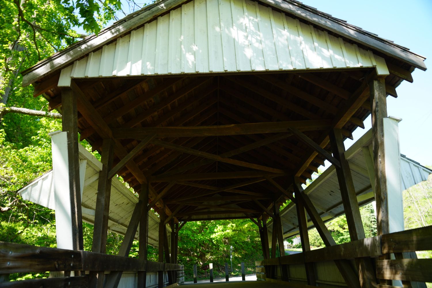 Interior view of a historic wooden covered bridge showing timber frame construction and green trees beyond.