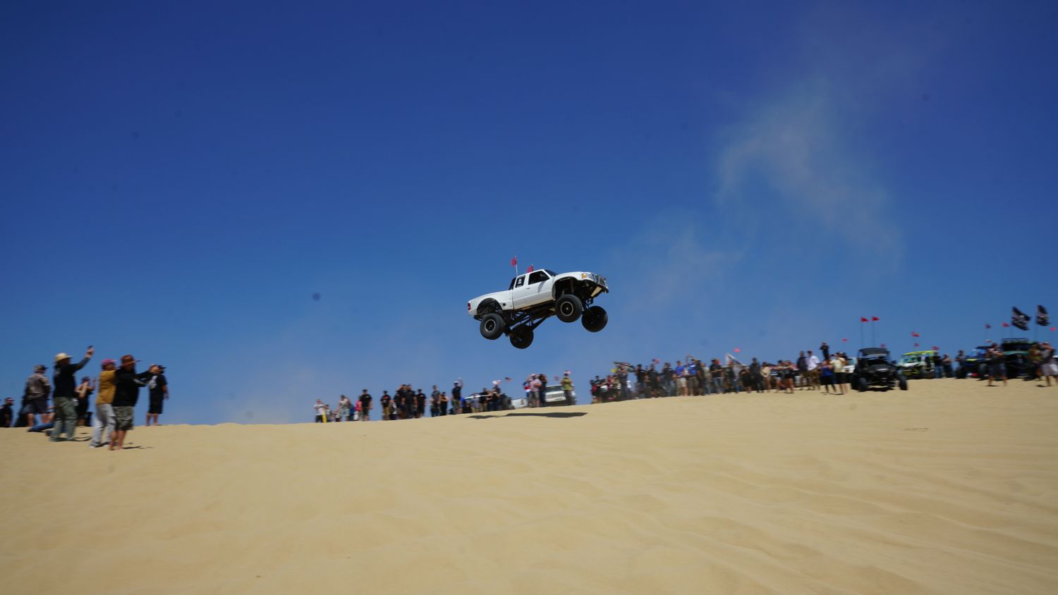 White monster truck catches big air over sandy dunes while crowds watch the off-road jumping event.