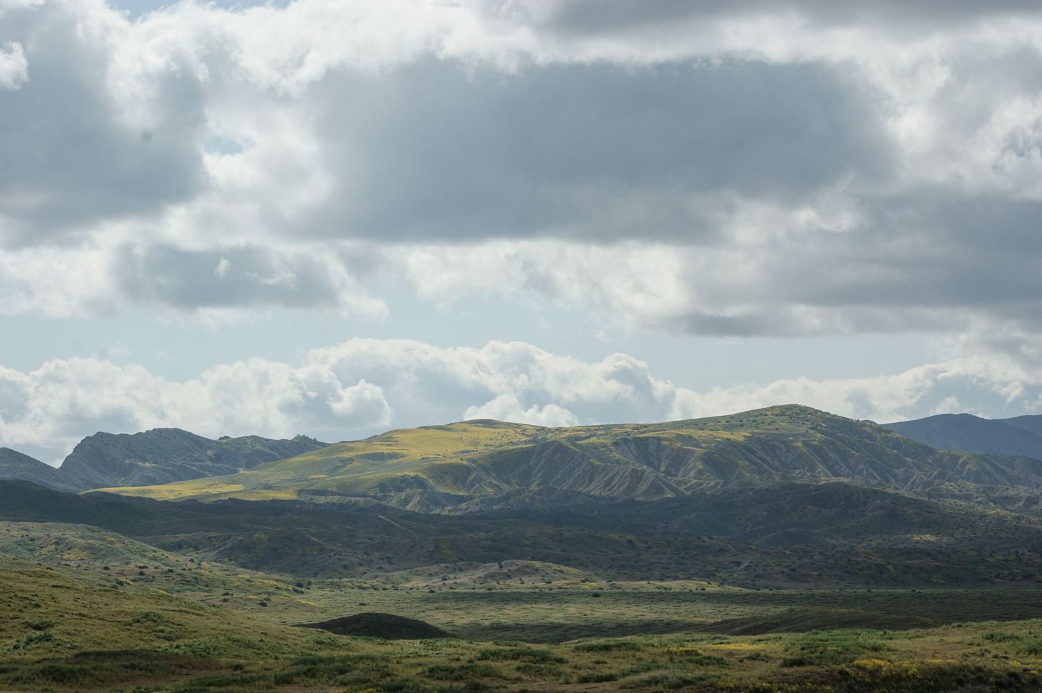 Expansive hilly landscape with green valleys and sunlit peaks under a dramatic cloudy sky.