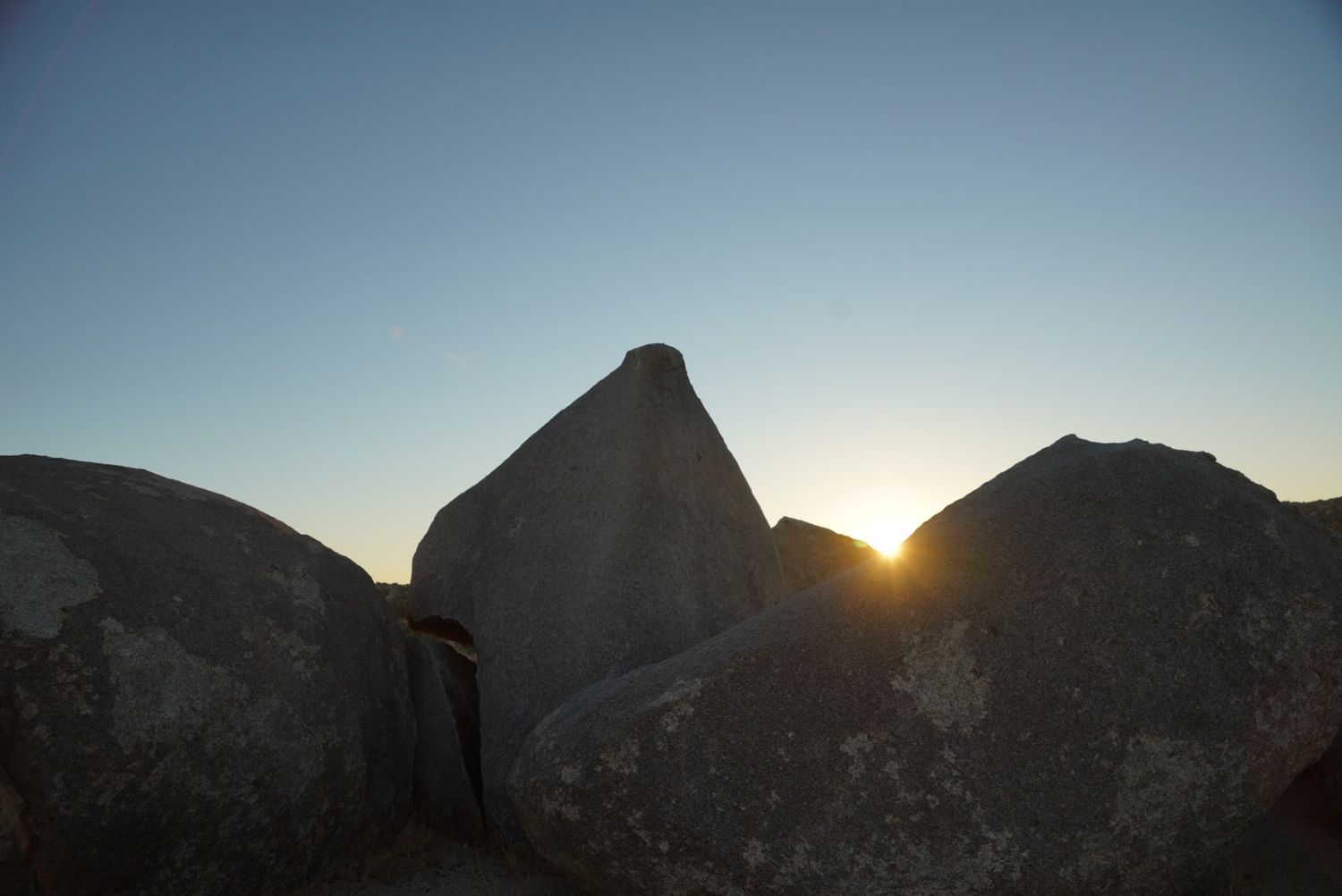 Sun setting behind large granite boulders creating a dramatic silhouette against a clear blue sky.