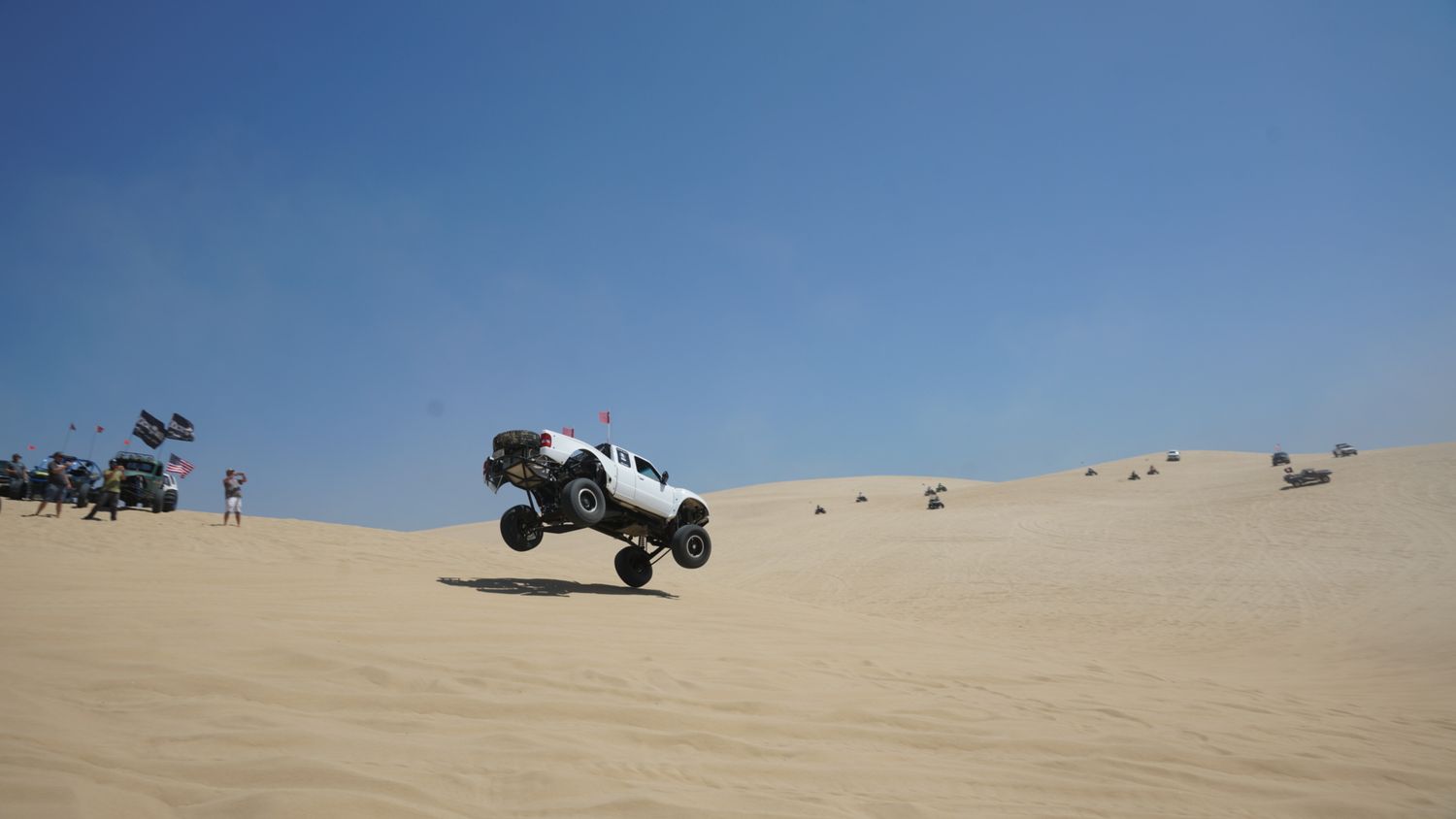 White off-road truck catches air over sand dunes at an outdoor desert racing event under clear blue skies.