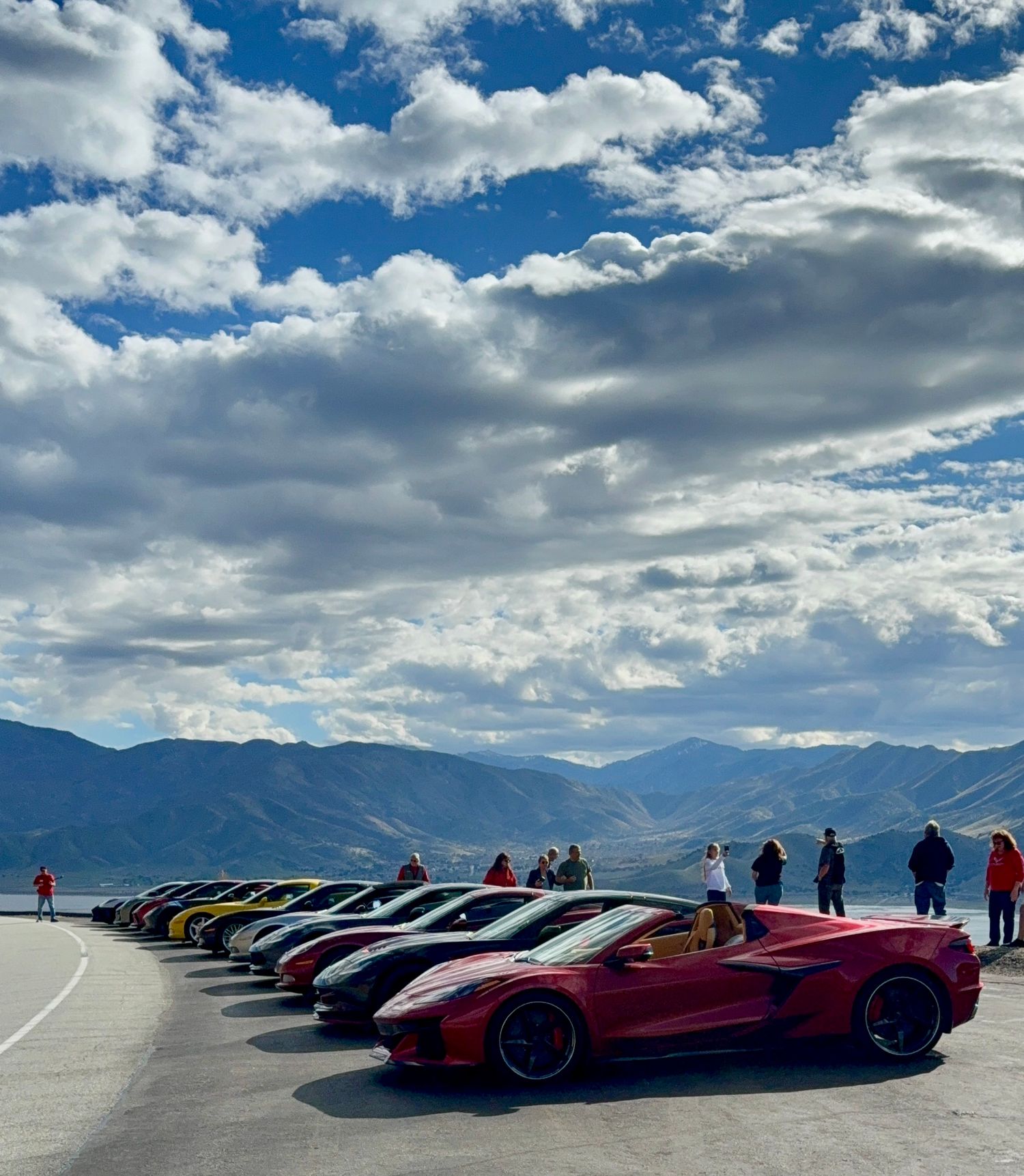 Row of colorful sports cars parked at a scenic mountain overlook under a blue cloudy sky.