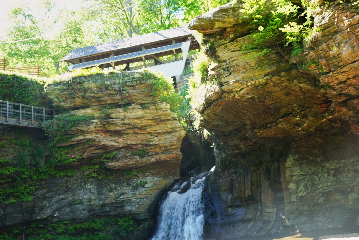 Scenic waterfall cascading down rocky cliffs beneath a wooden covered bridge in a lush gorge.