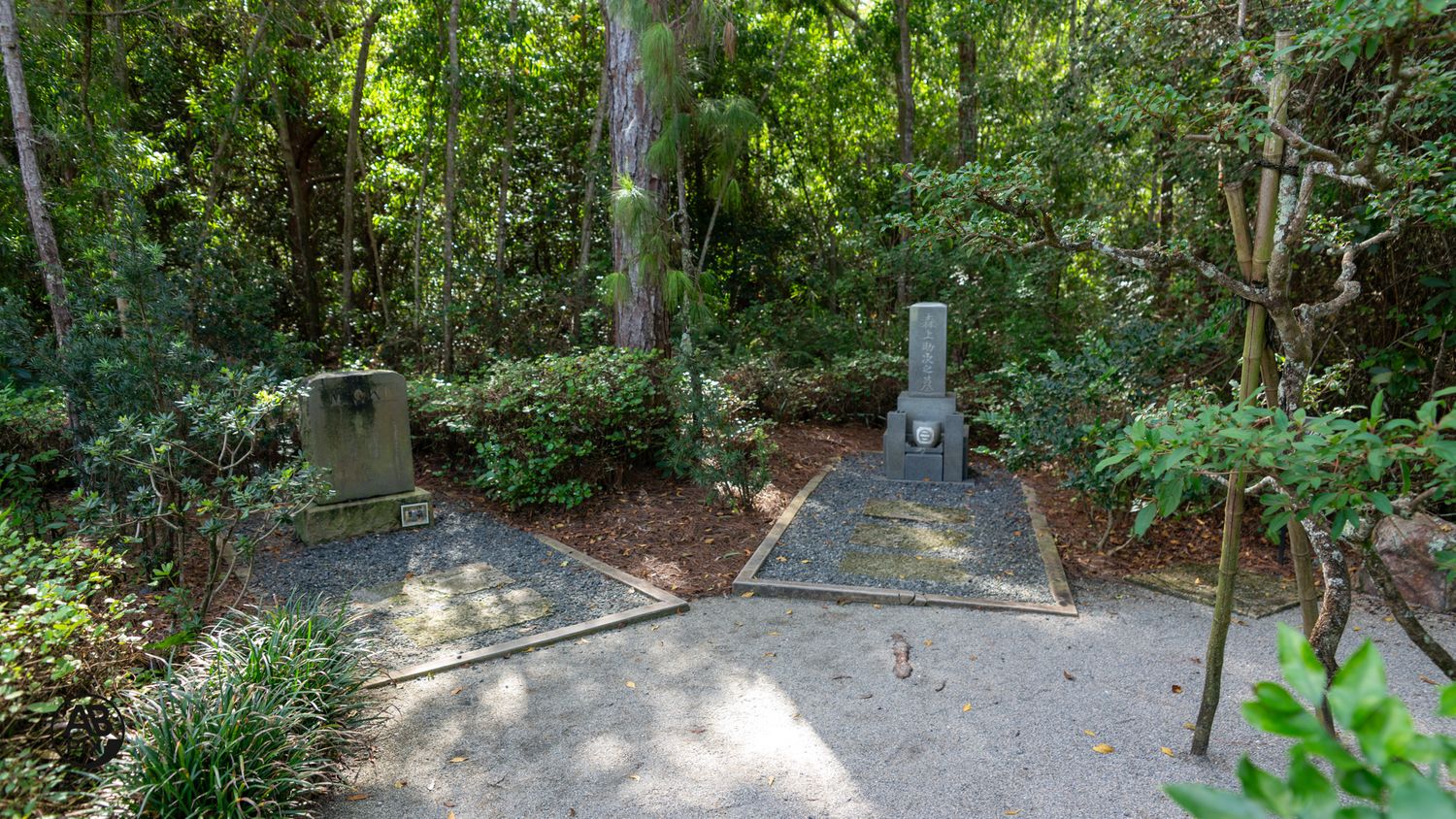Two stone grave markers surrounded by tall trees in a serene, wooded Japanese garden setting.