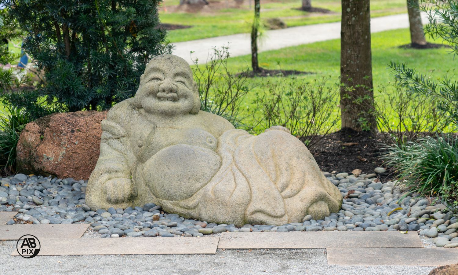 Smiling stone Buddha statue resting on river rocks in a garden landscape.