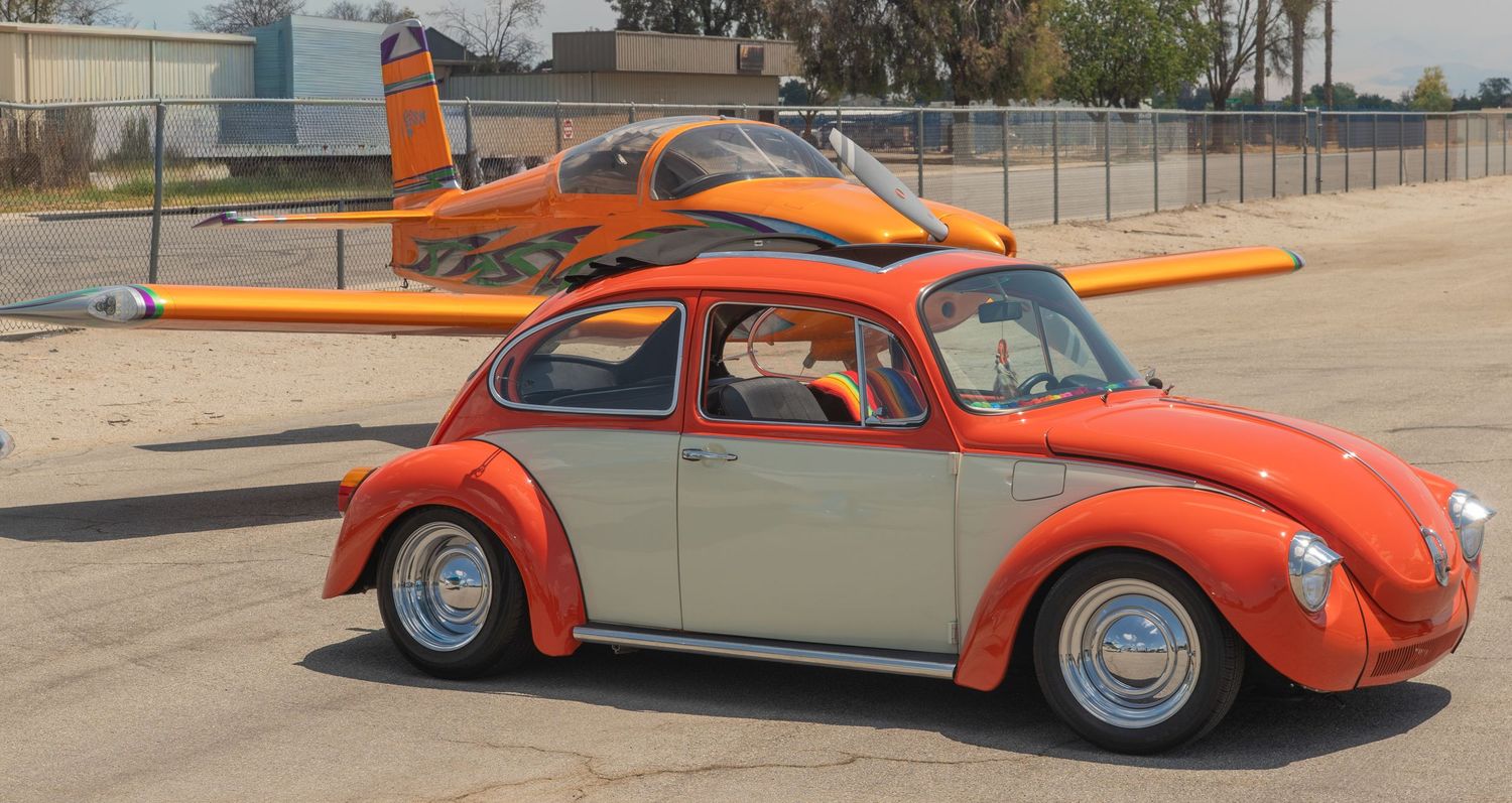 Vintage orange and white VW Beetle parked on tarmac with a matching orange jet trainer aircraft in the background.
