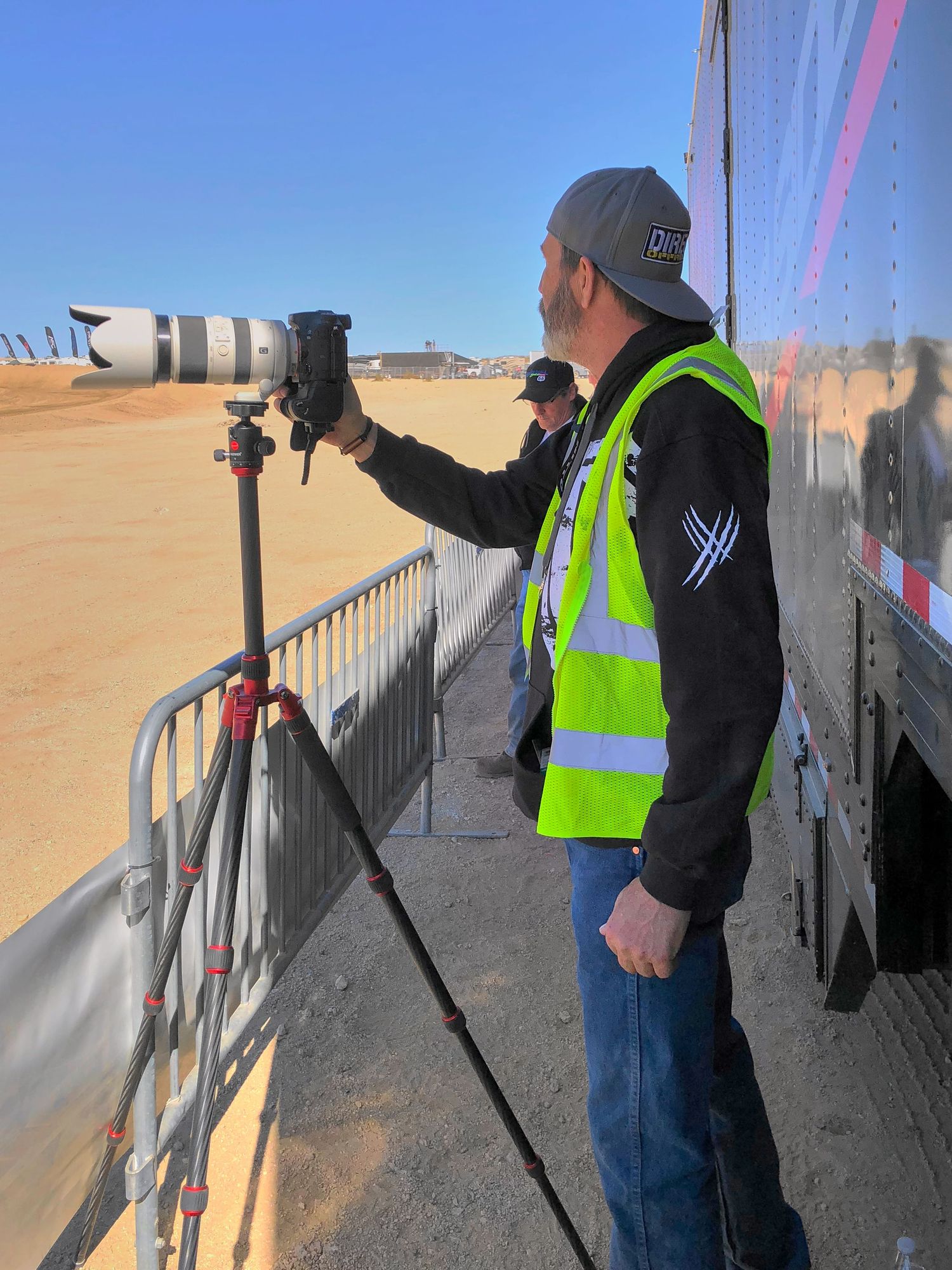 Photographer in neon yellow safety vest uses a telephoto lens on a tripod at an outdoor racing event.