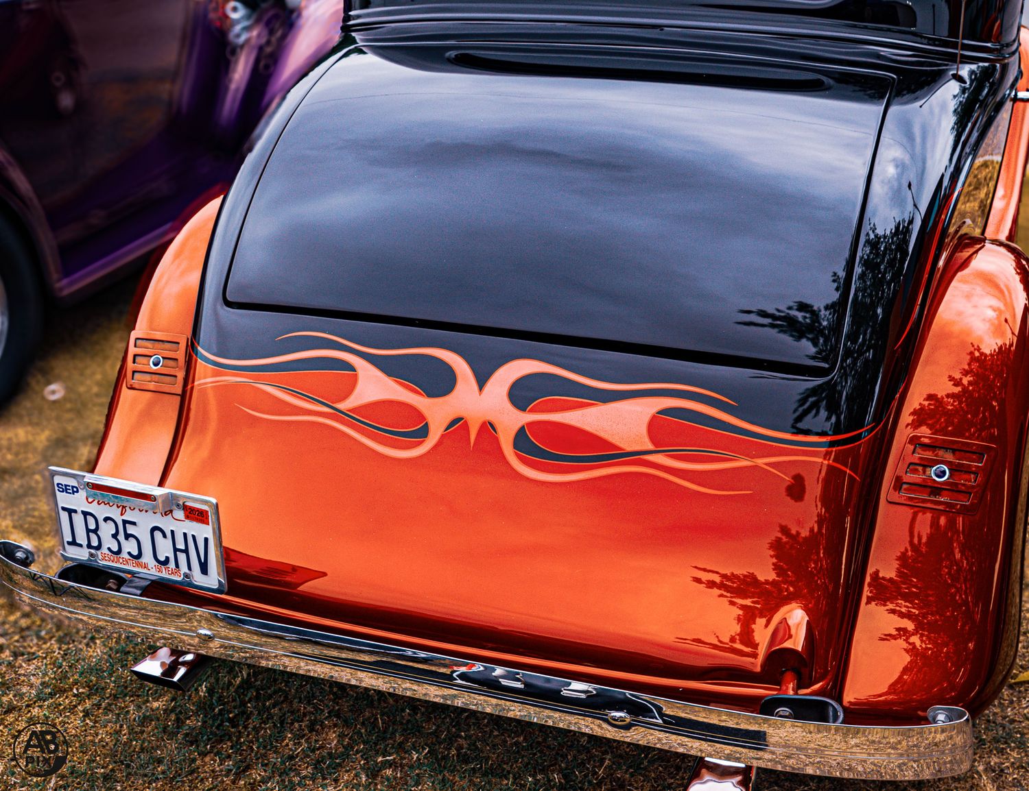 Orange classic hot rod with flame graphics on trunk lid and vintage license plate displayed outdoors.
