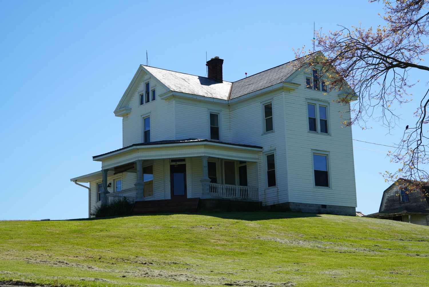 White two-story farmhouse with wrap-around porch and chimney sitting atop a green hill under blue sky.