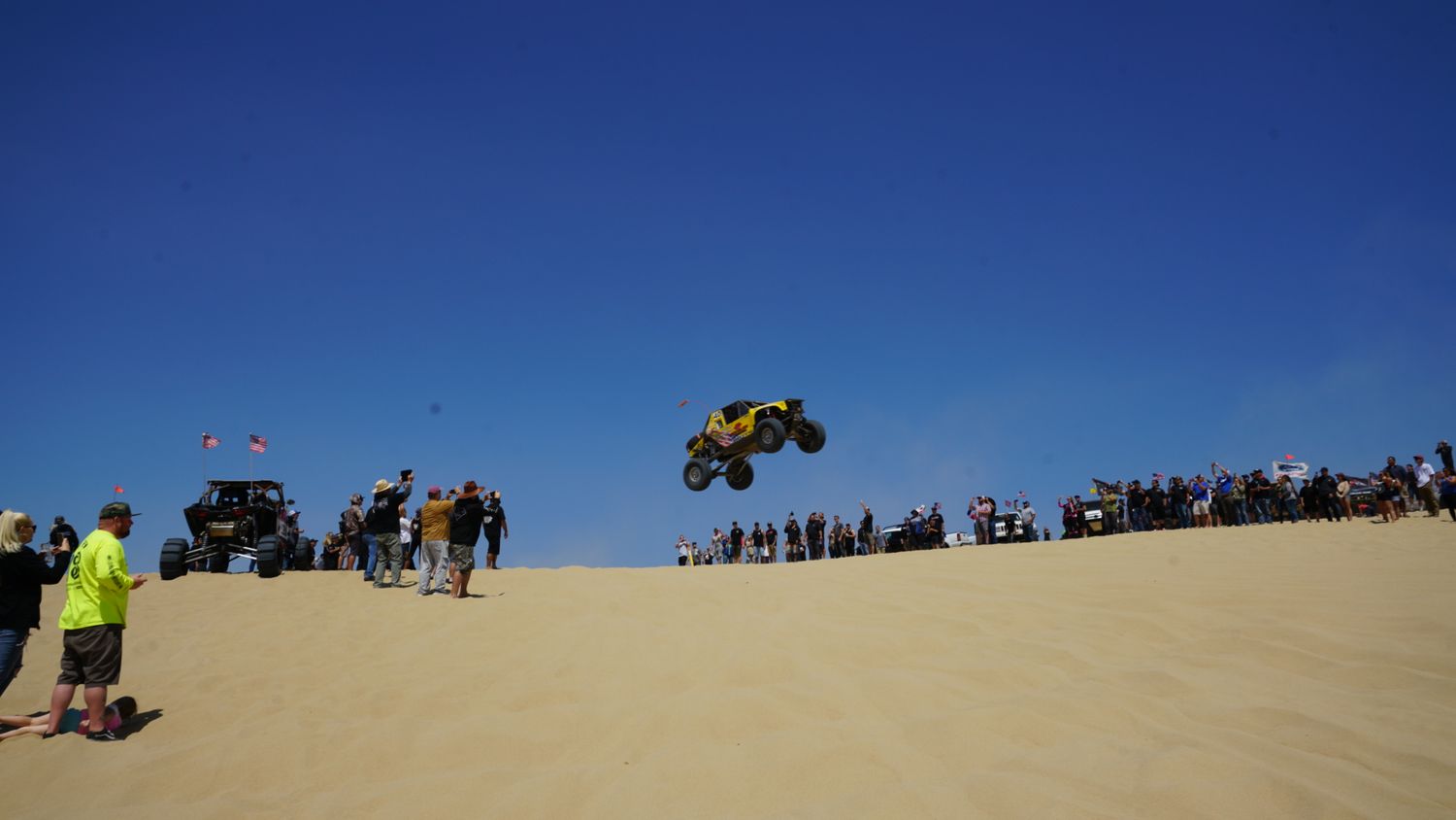 Yellow off-road buggy soars above sand dunes in front of a large crowd at a desert racing event.
