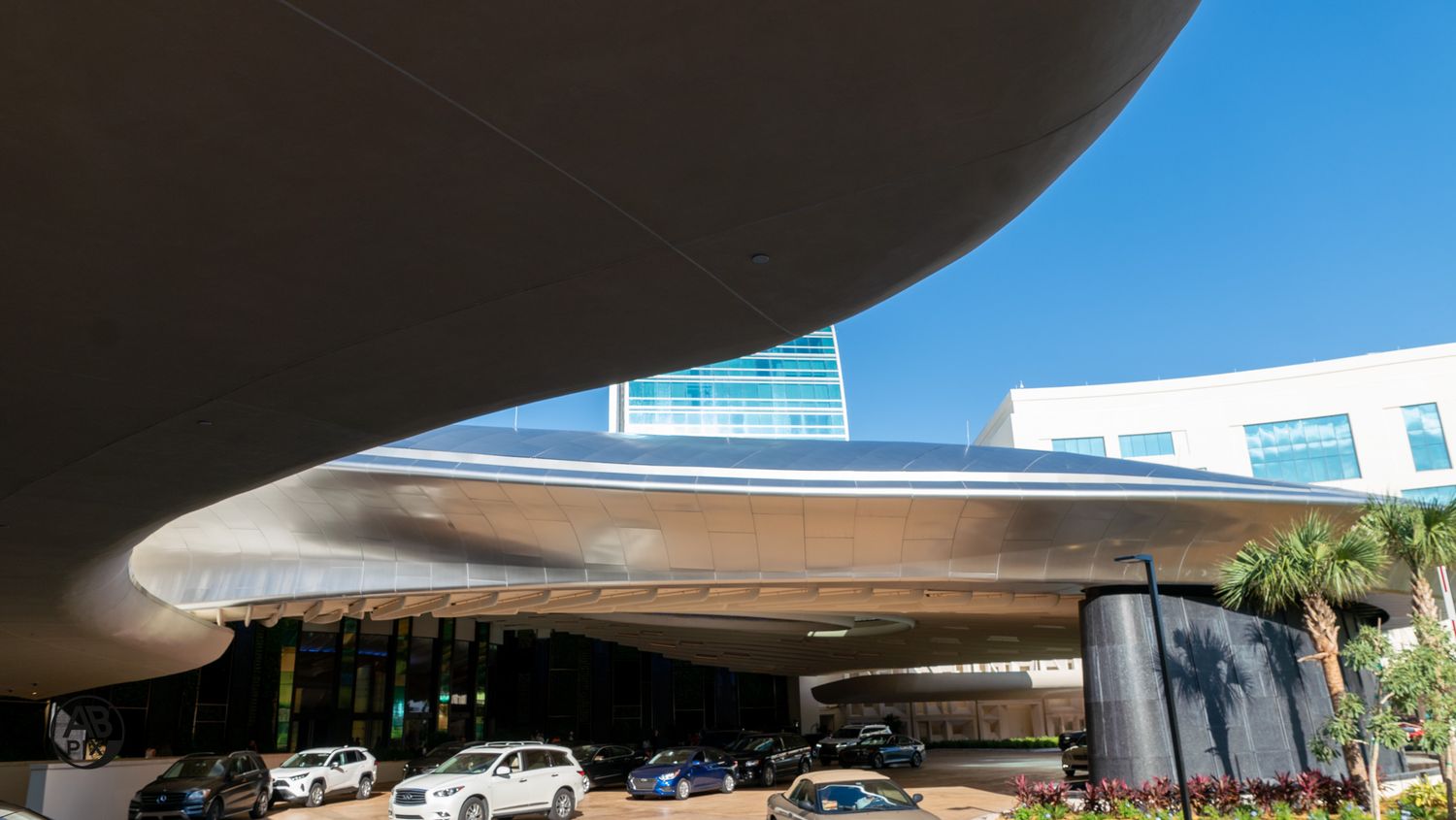 Sweeping curved modern architectural canopy over hotel entrance with palm trees and parked cars.