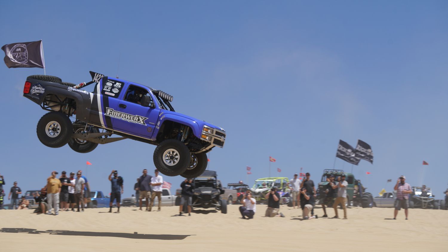 A blue off-road trophy truck catching massive air over sand dunes at an outdoor motorsport event.