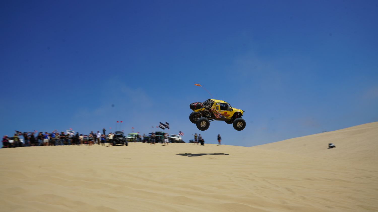 Yellow dune buggy catching massive air over sandy dunes with crowd of spectators watching the off-road stunt.