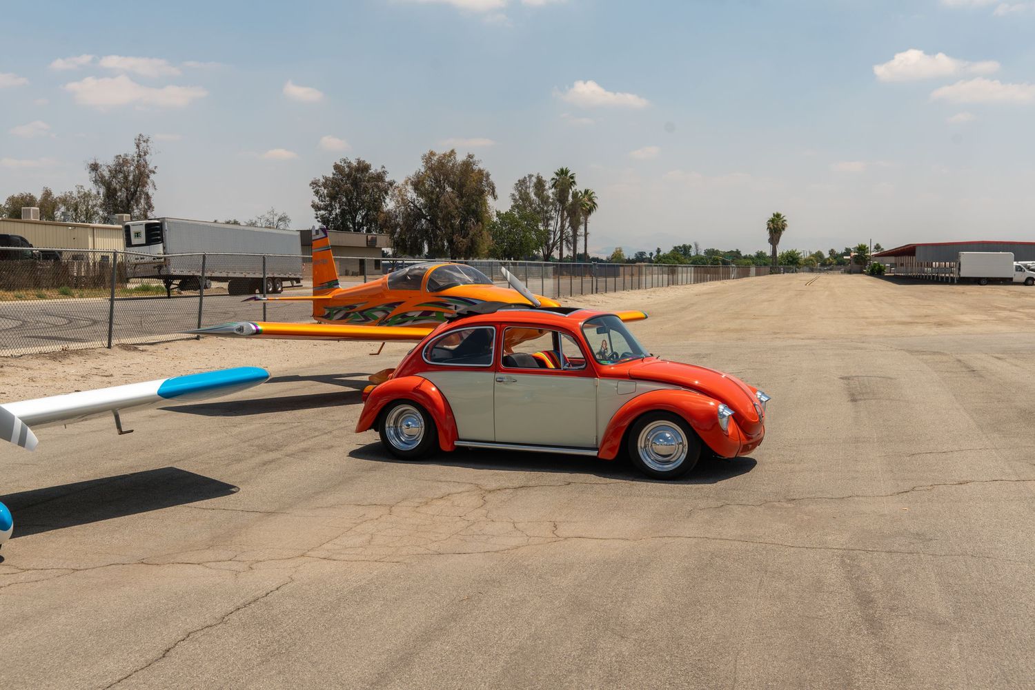 A classic red and white VW Beetle parked on an airstrip next to a small orange and blue airplane.