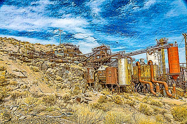 Abandoned rusty mining facility with industrial equipment on a rocky desert hillside under blue sky.