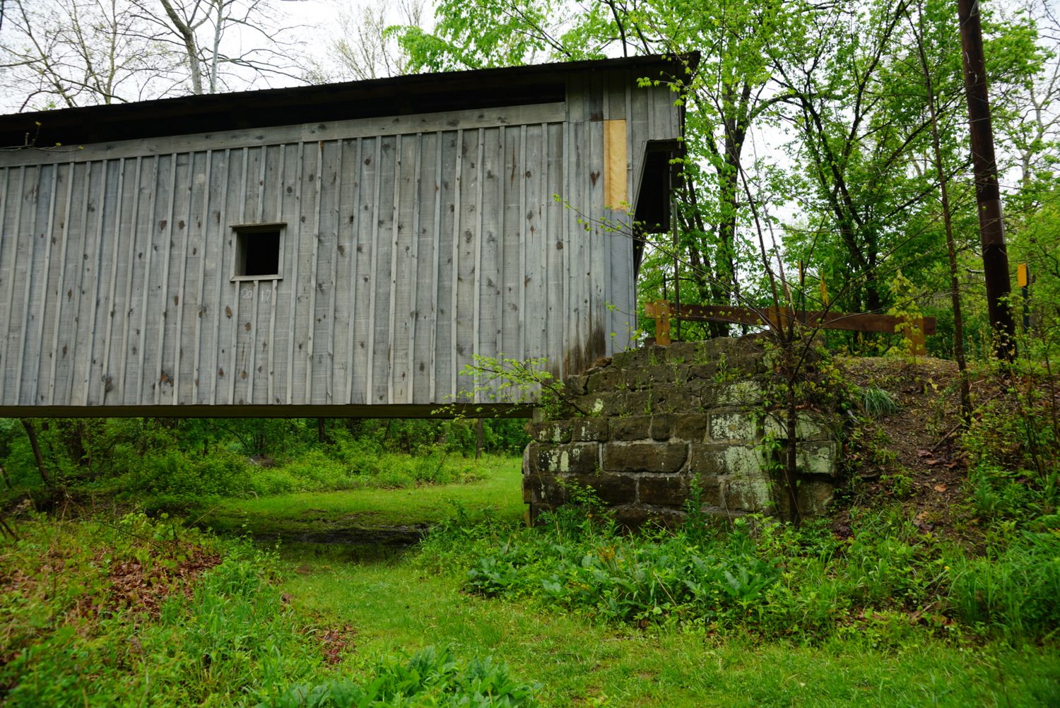 The weathered gray wooden side of an old covered bridge rests on a mossy stone foundation in woods.