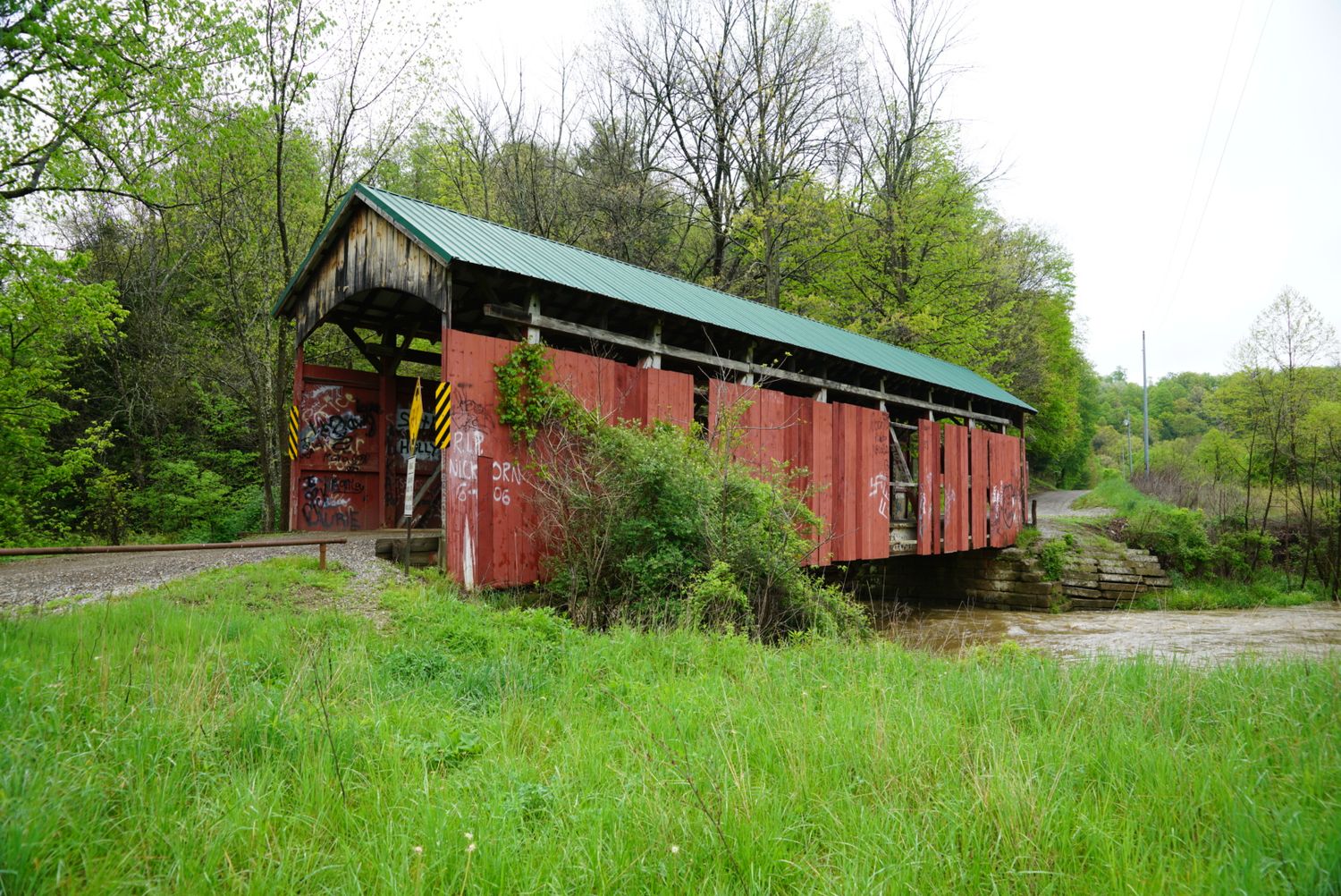 A historic red covered bridge with a green roof sits in a grassy field surrounded by spring trees.