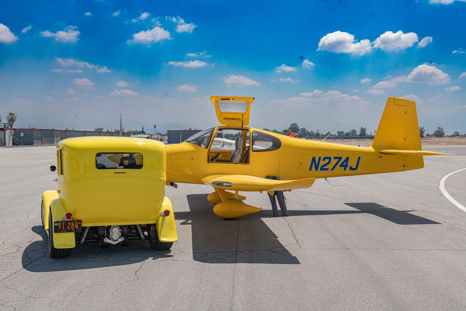 Bright yellow small aircraft N274J parked on airport tarmac beside a matching yellow vintage car under blue sky.