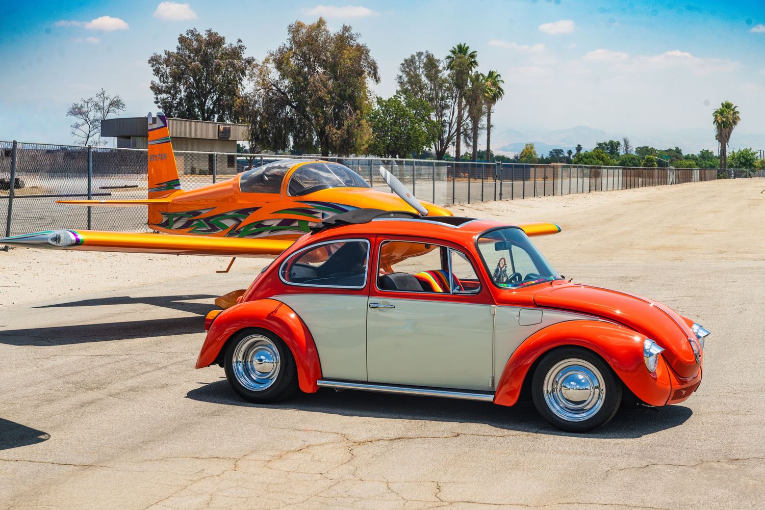 Orange and white custom VW Beetle parked on tarmac beside a yellow aircraft at a sunny airfield.