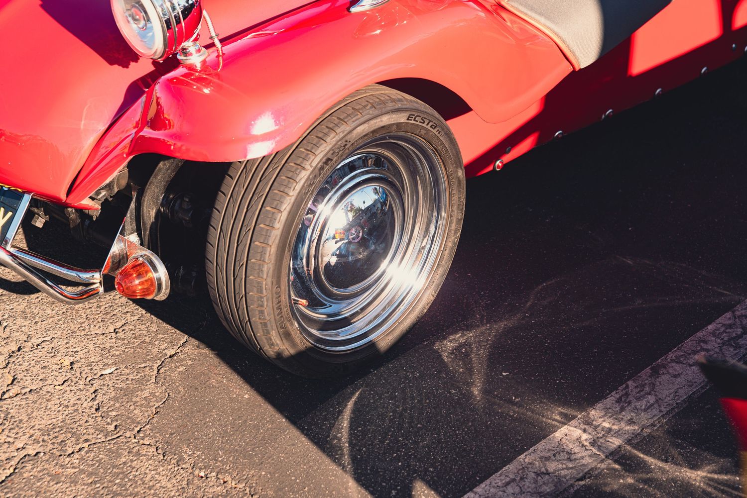 Close-up of a classic red vintage car's chrome wheel and tire on asphalt at an outdoor car show.