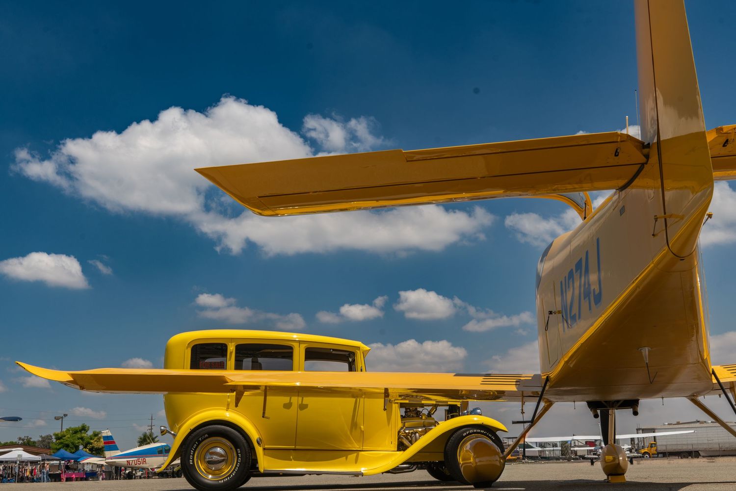 Bright yellow hot rod car parked beneath the wing of a yellow airplane under blue skies.
