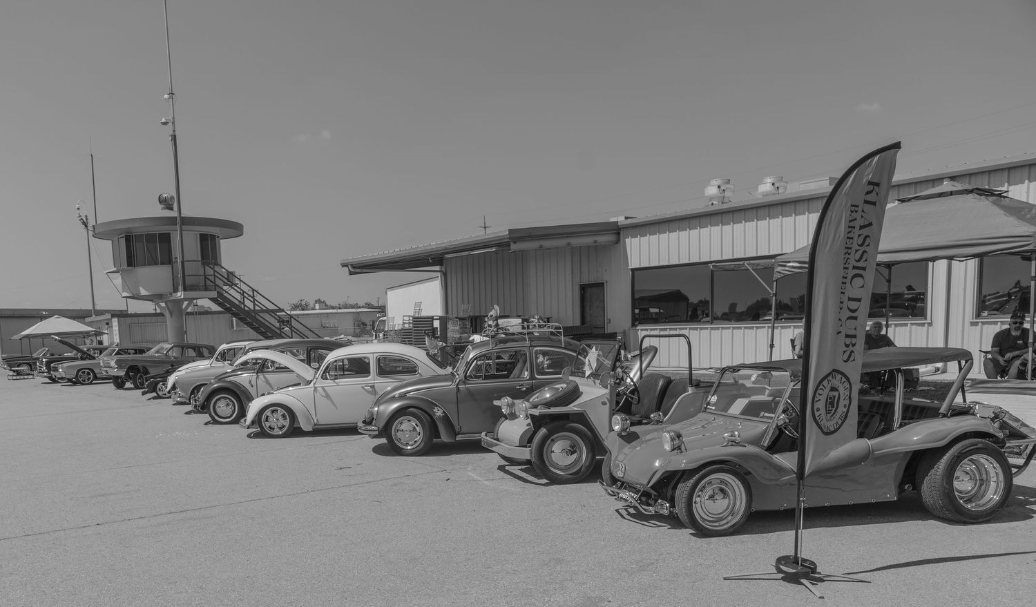 Vintage VW Beetles lined up at an airfield garage with a promotional banner in this classic black and white photo.