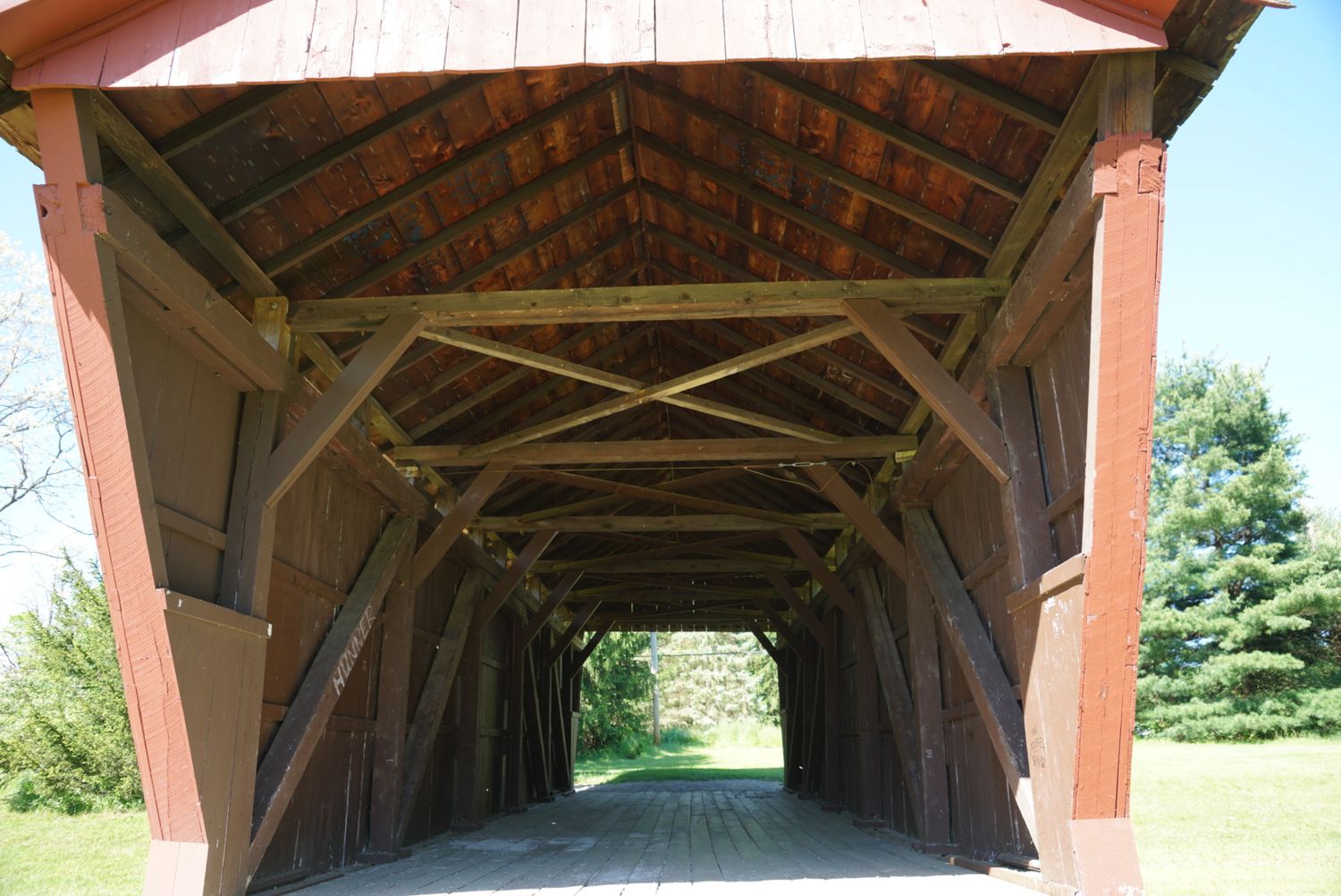 Interior of a red painted historic covered bridge with wooden truss beams and open ends.