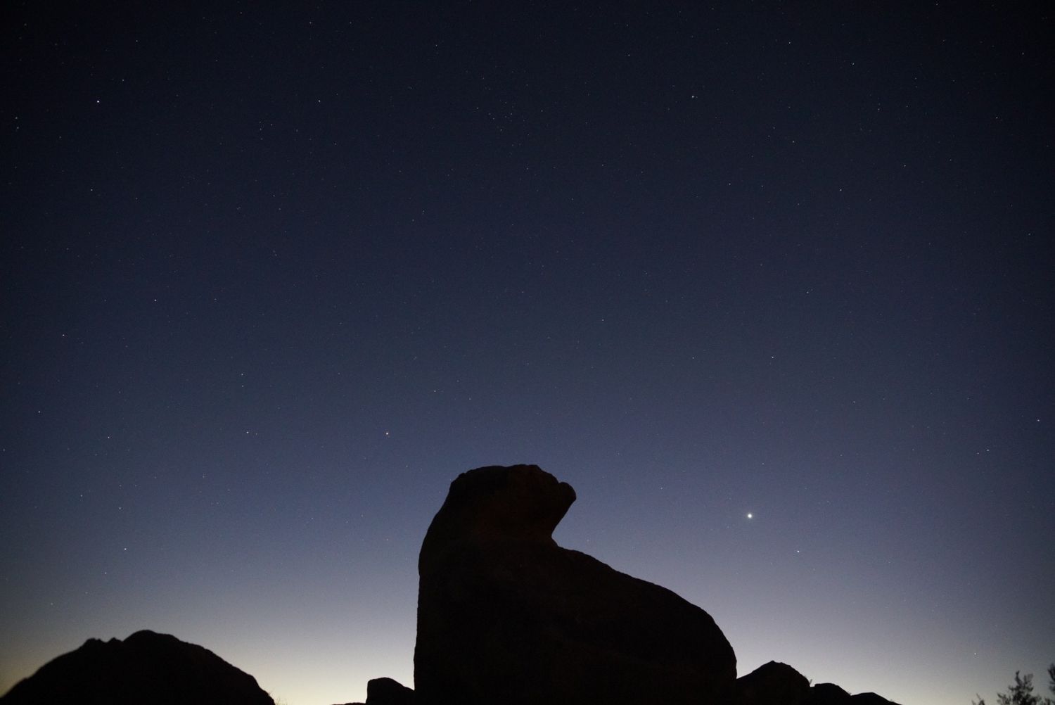 Silhouette of large desert rock formation against starry twilight sky.