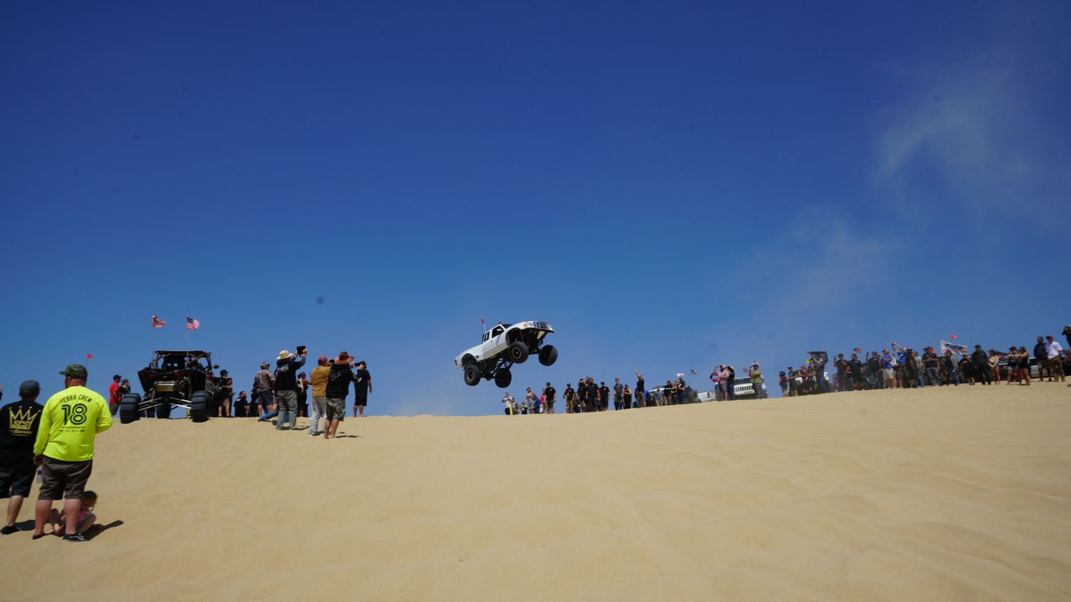 White off-road truck launches off a sand dune in front of spectators at a desert motorsport event.