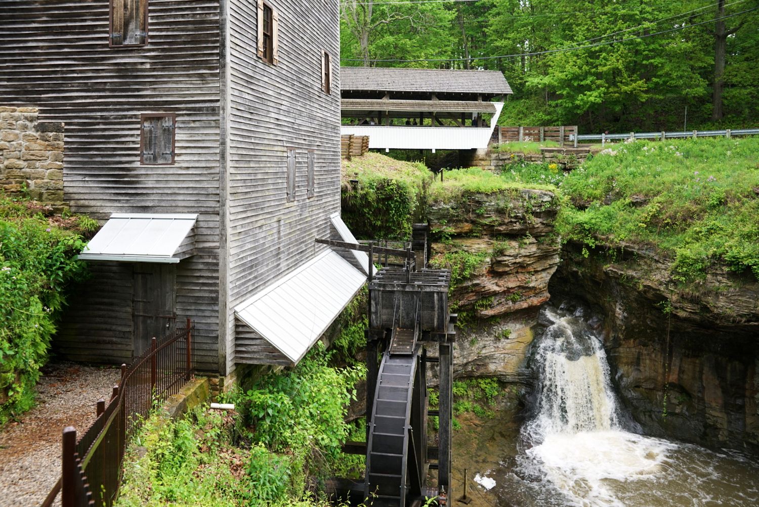 Historic grist mill with wooden water wheel beside a rocky waterfall and covered bridge surrounded by lush green trees.