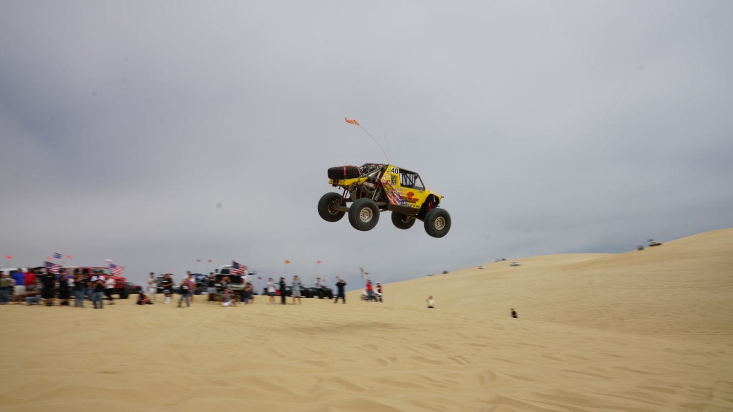 Yellow off-road buggy catching big air over sand dunes with large crowd of spectators watching below.