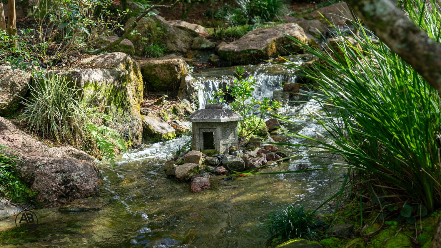 Small stone pagoda lantern beside a gentle waterfall stream among moss-covered rocks in a Japanese garden.