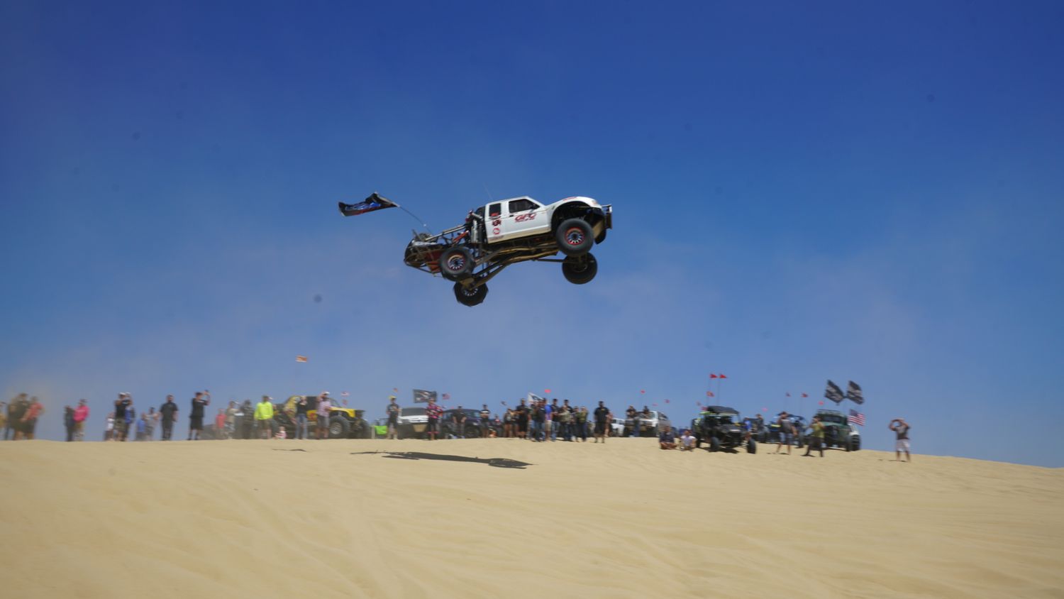 White 4x4 trophy truck catches massive air over sand dunes at a crowded off-road racing event.