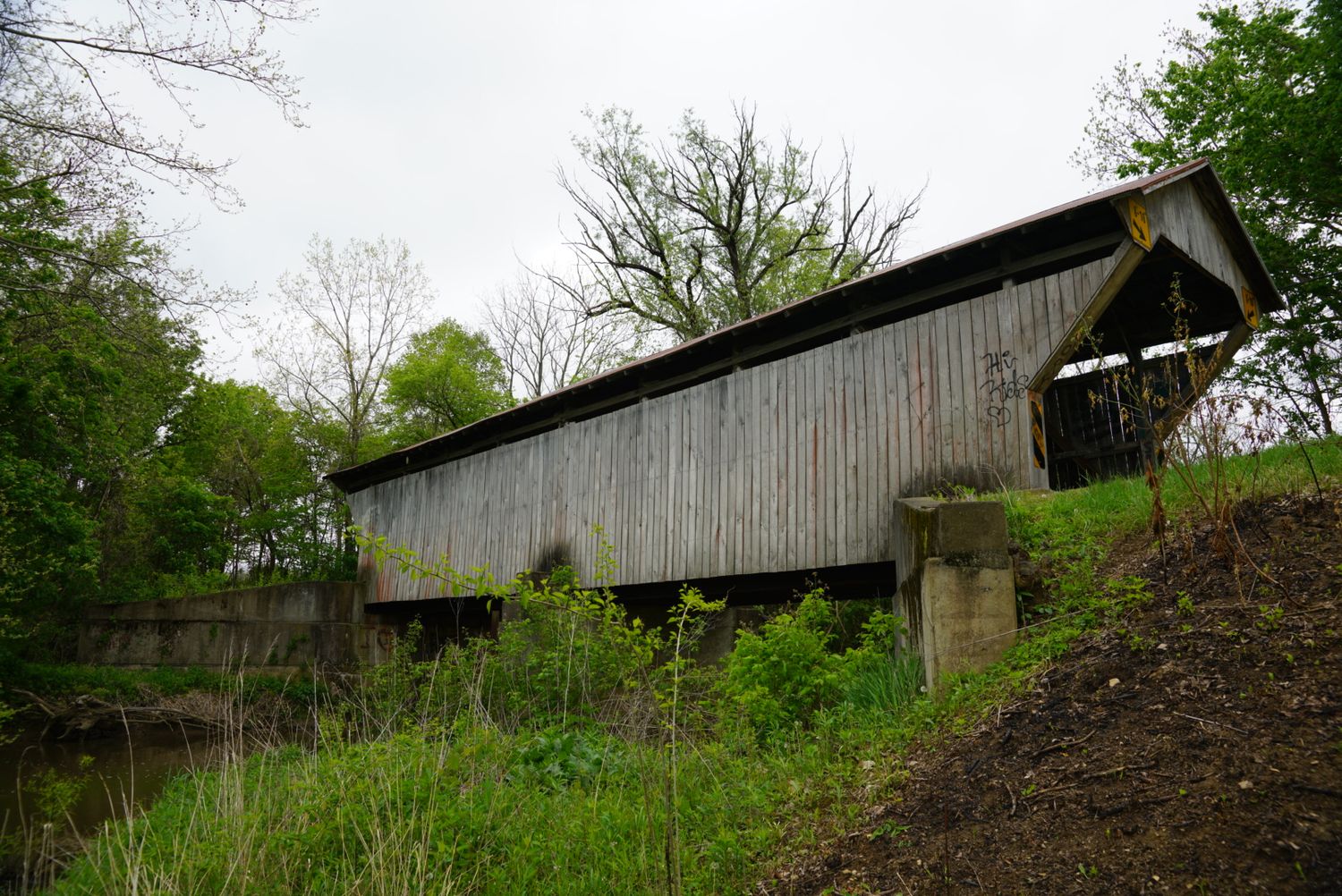 A weathered wooden covered bridge spans a creek on a cloudy spring day with green vegetation.