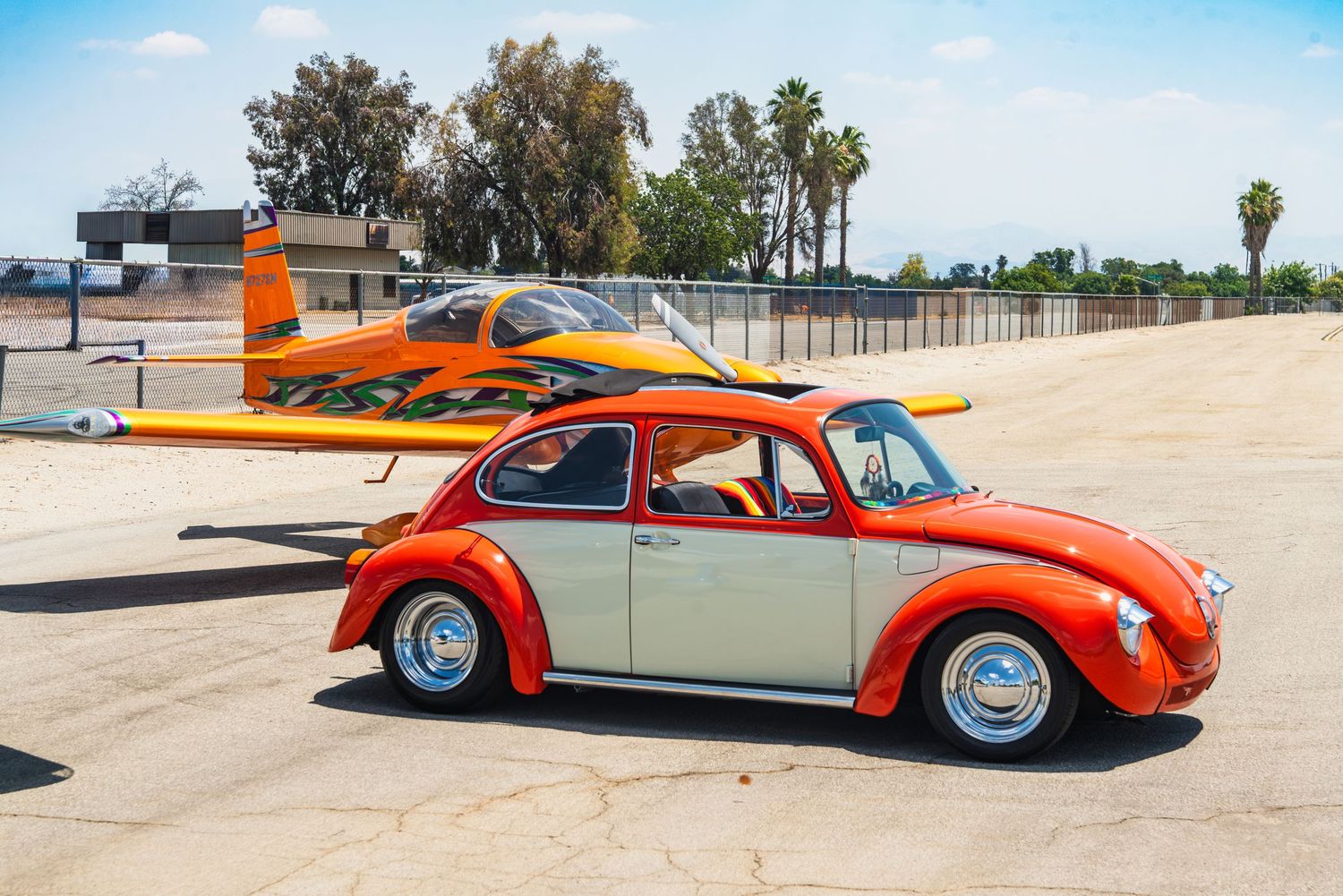 Orange and white custom VW Beetle parked in front of an orange and yellow airplane on a tarmac.