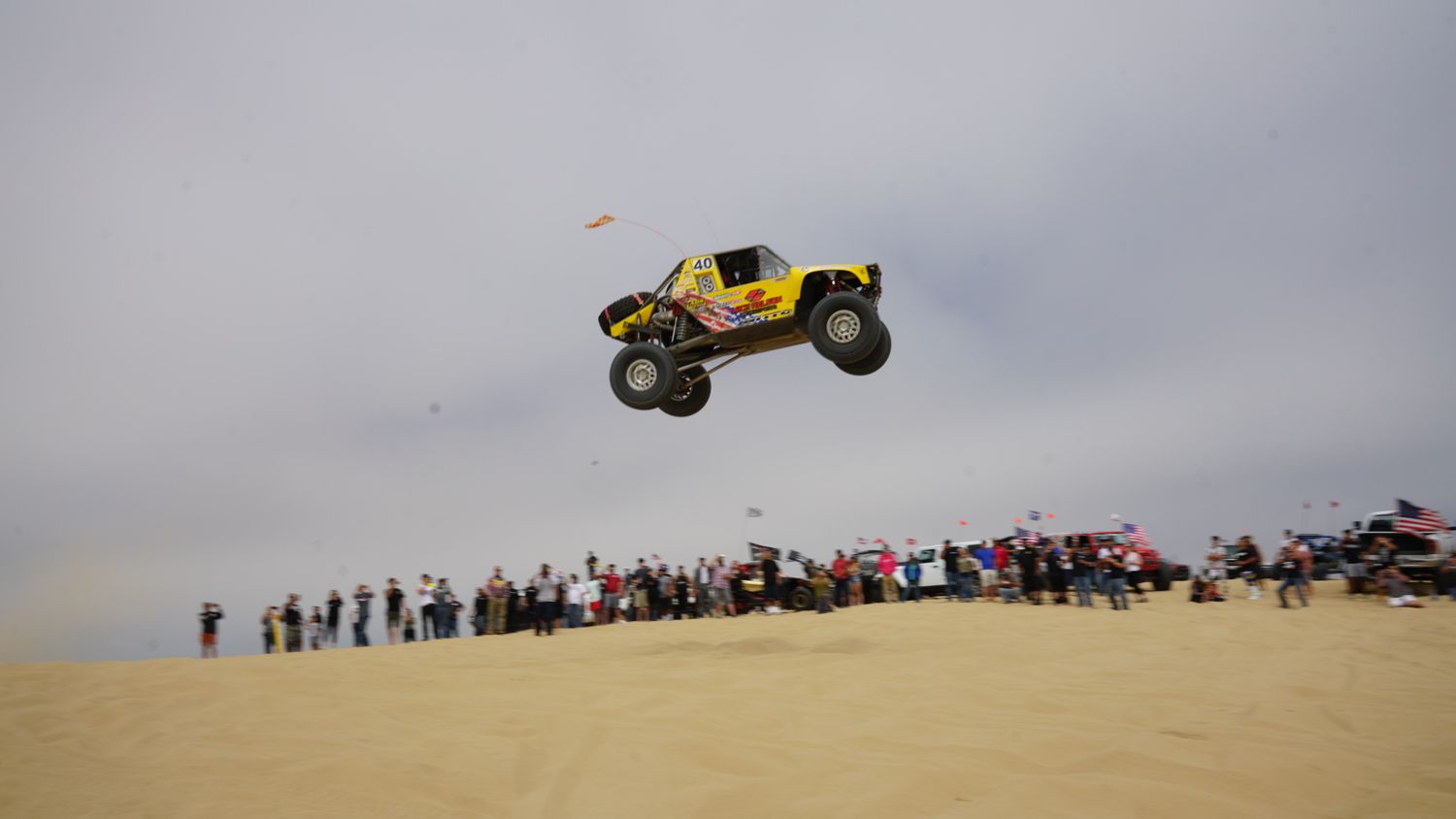 Yellow off-road buggy soaring high above sand dunes while a large crowd of spectators watches the jump.