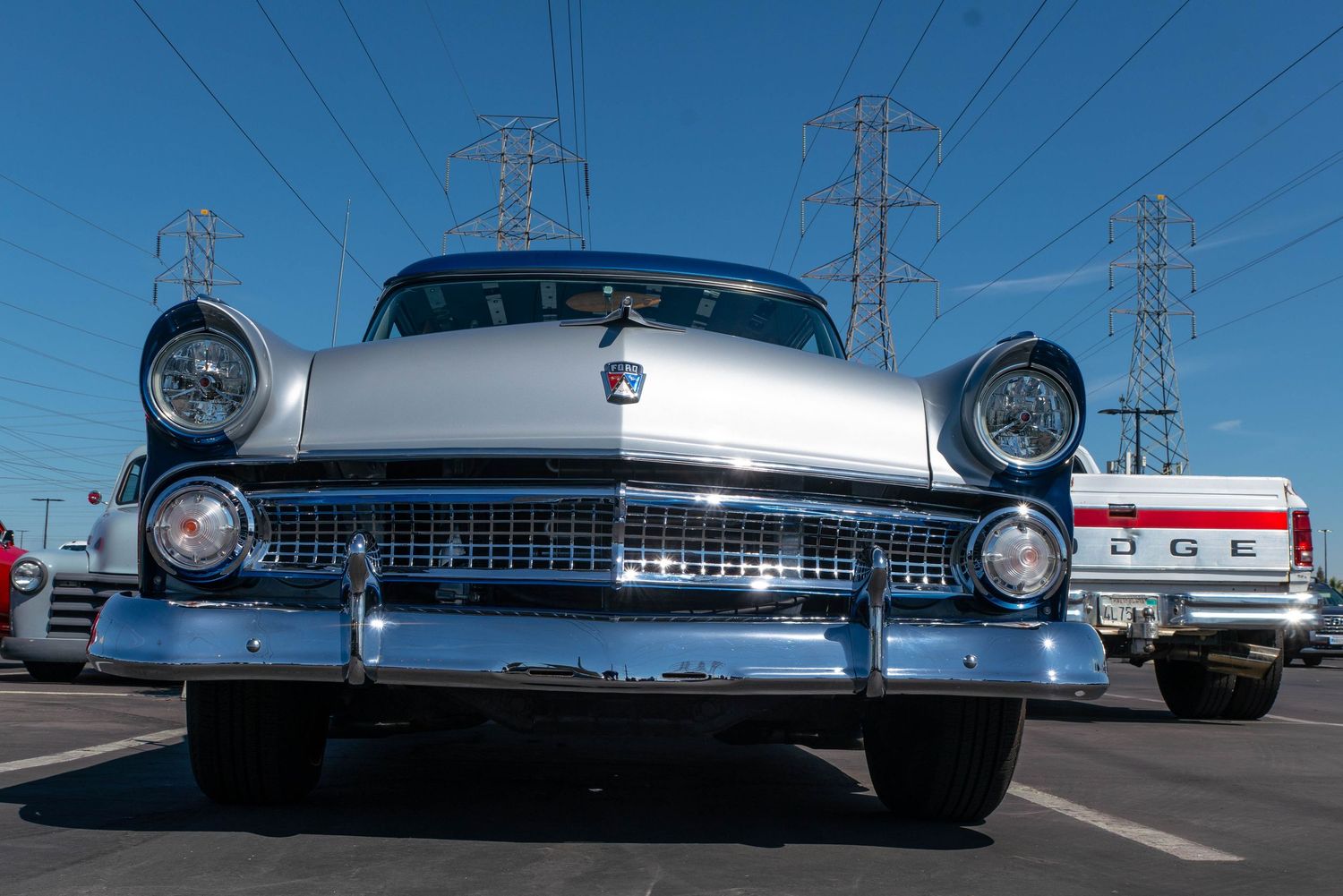 Front view of a classic 1950s Ford car with chrome grille, parked near power lines with a Dodge truck in background.