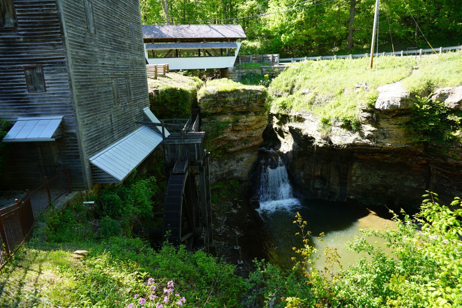 A historic grist mill with a water wheel sits beside a rocky waterfall cliff surrounded by green trees.