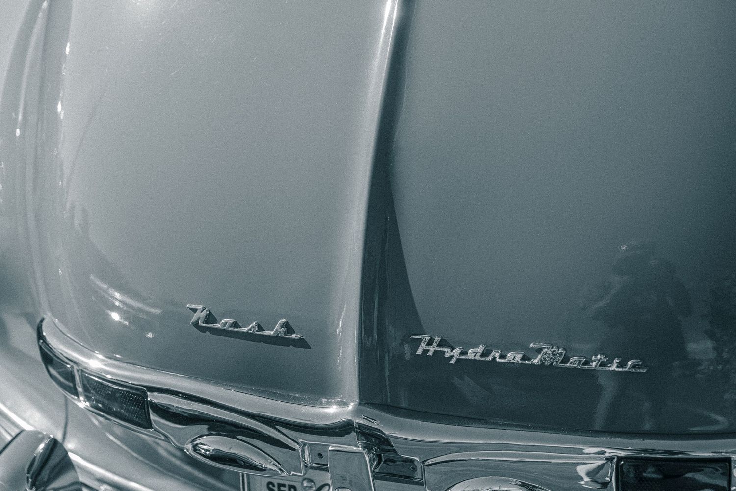 Black and white close-up of a classic car hood featuring chrome Hydra-Matic and Nash emblems.