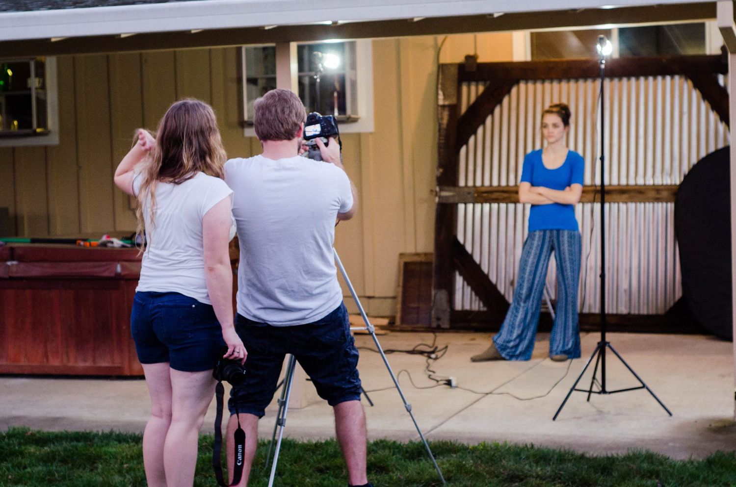 Two people filming a woman posing under studio lights outside a barn at dusk.