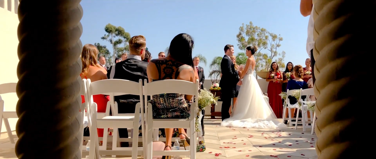 Outdoor wedding ceremony with guests seated in white chairs as bride walks down the aisle.
