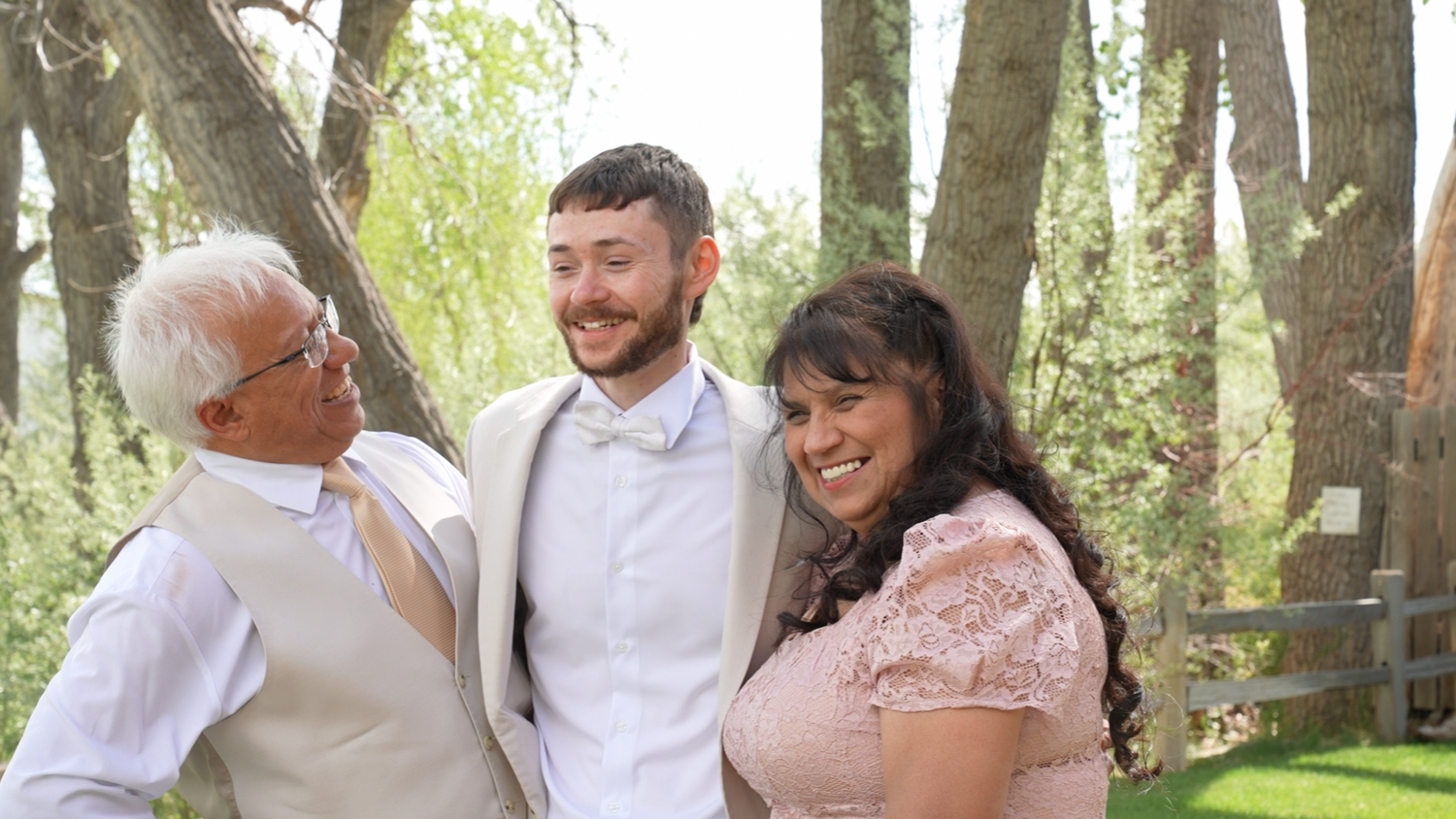 Three people smiling outdoors at a wedding, with groom in white suit flanked by family members among trees.