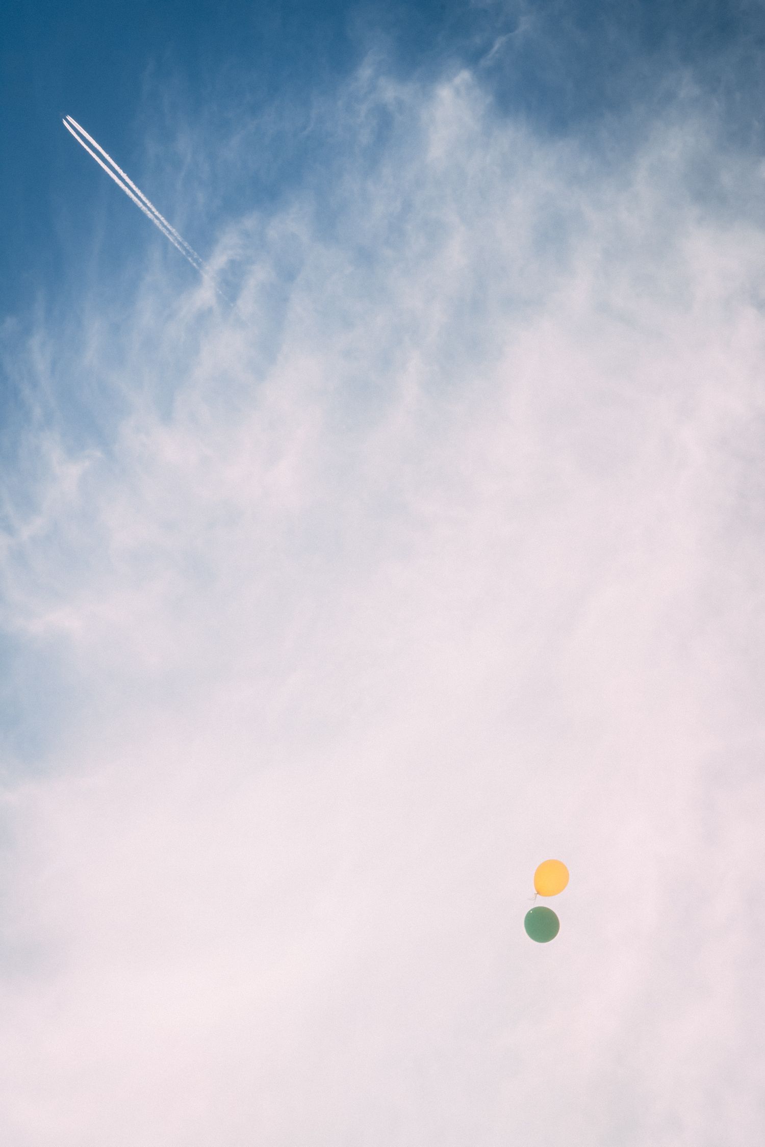 Blue sky with wispy clouds, two colorful balloons floating mid-air, and an airplane contrail streaking across the upper left.
