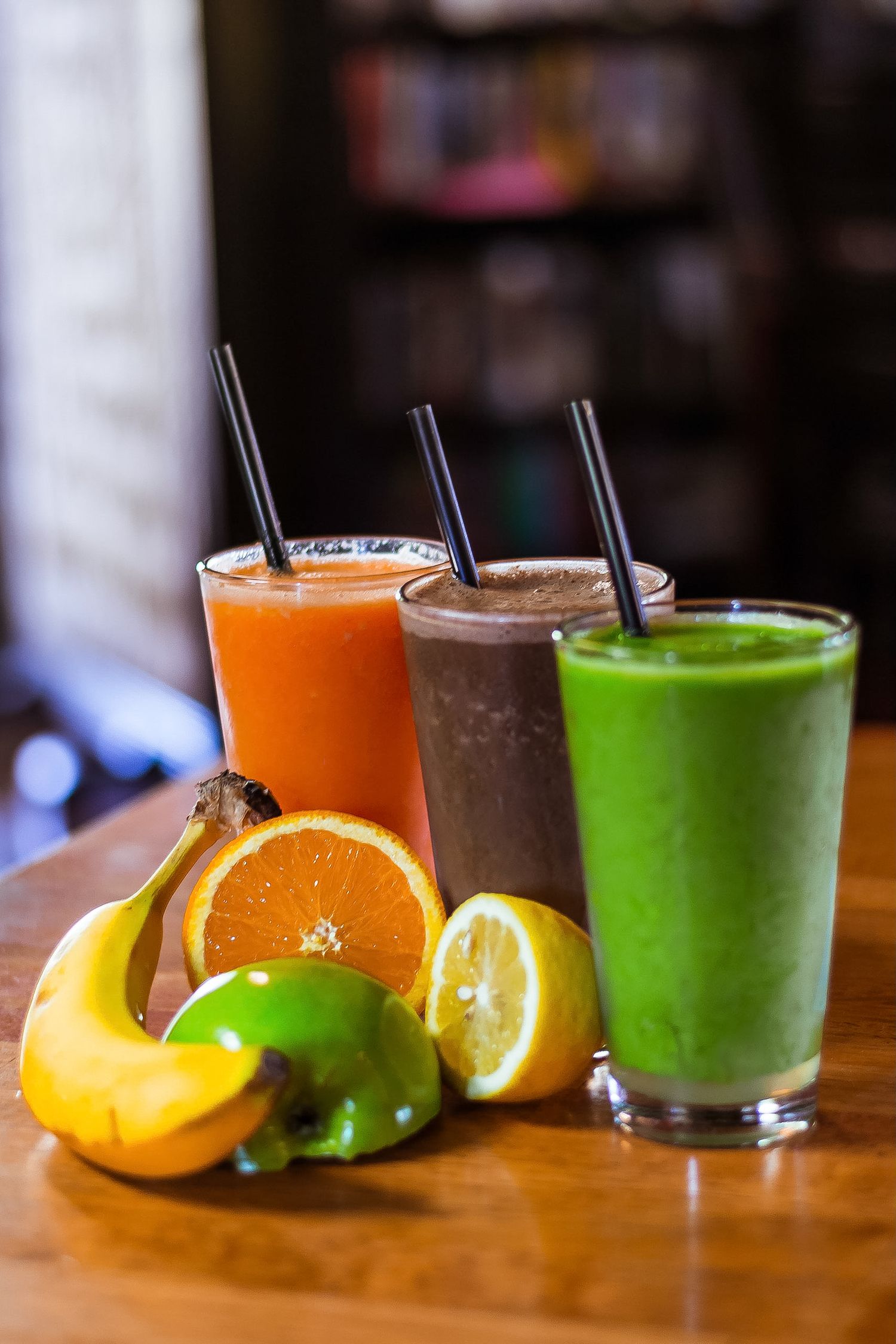Three colorful fruit smoothies with straws beside fresh banana, orange, lime, and lemon on wood table.