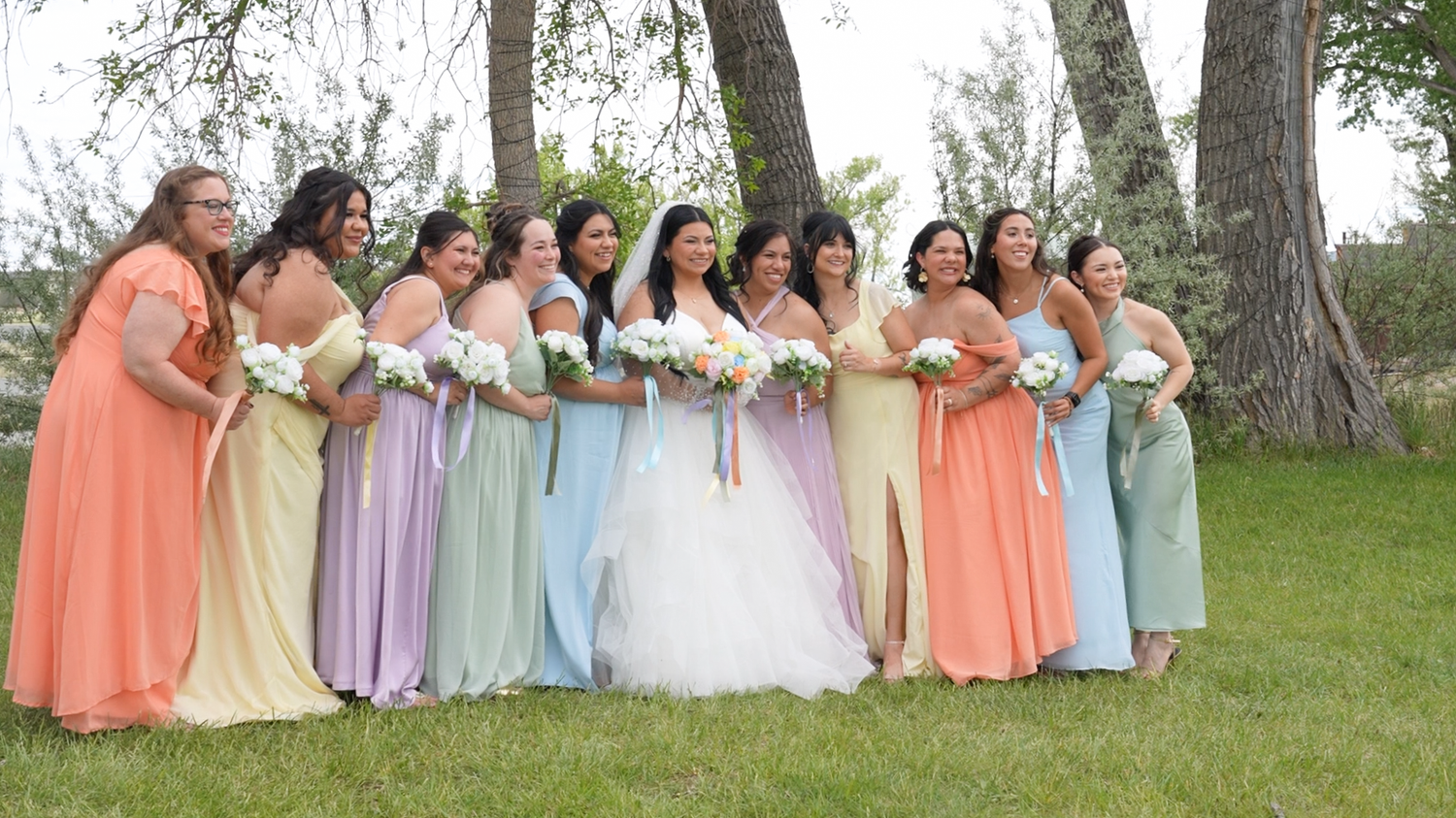Bride in white gown poses with bridesmaids in colorful pastel dresses holding bouquets outdoors near trees.