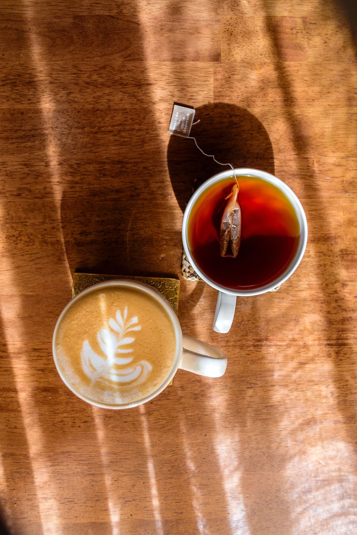 Overhead view of latte art coffee and tea with steeping bag on wooden table in warm sunlight.