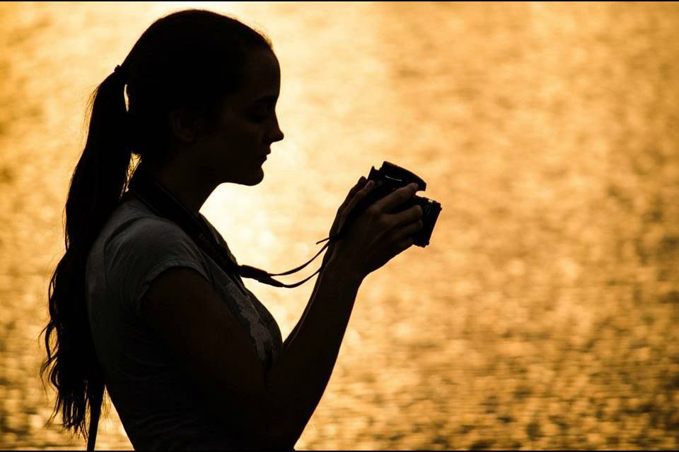 Silhouette of a woman holding a camera against a golden sunset reflected on water.