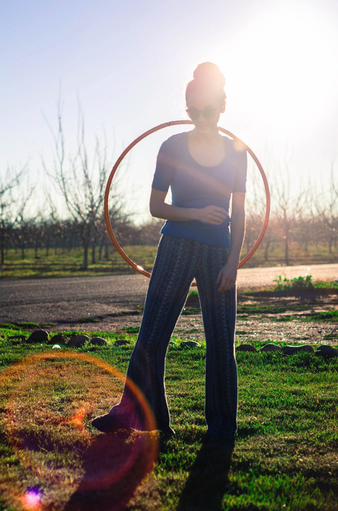 Woman holding a hula hoop outdoors in sunlight with lens flare in an orchard setting.