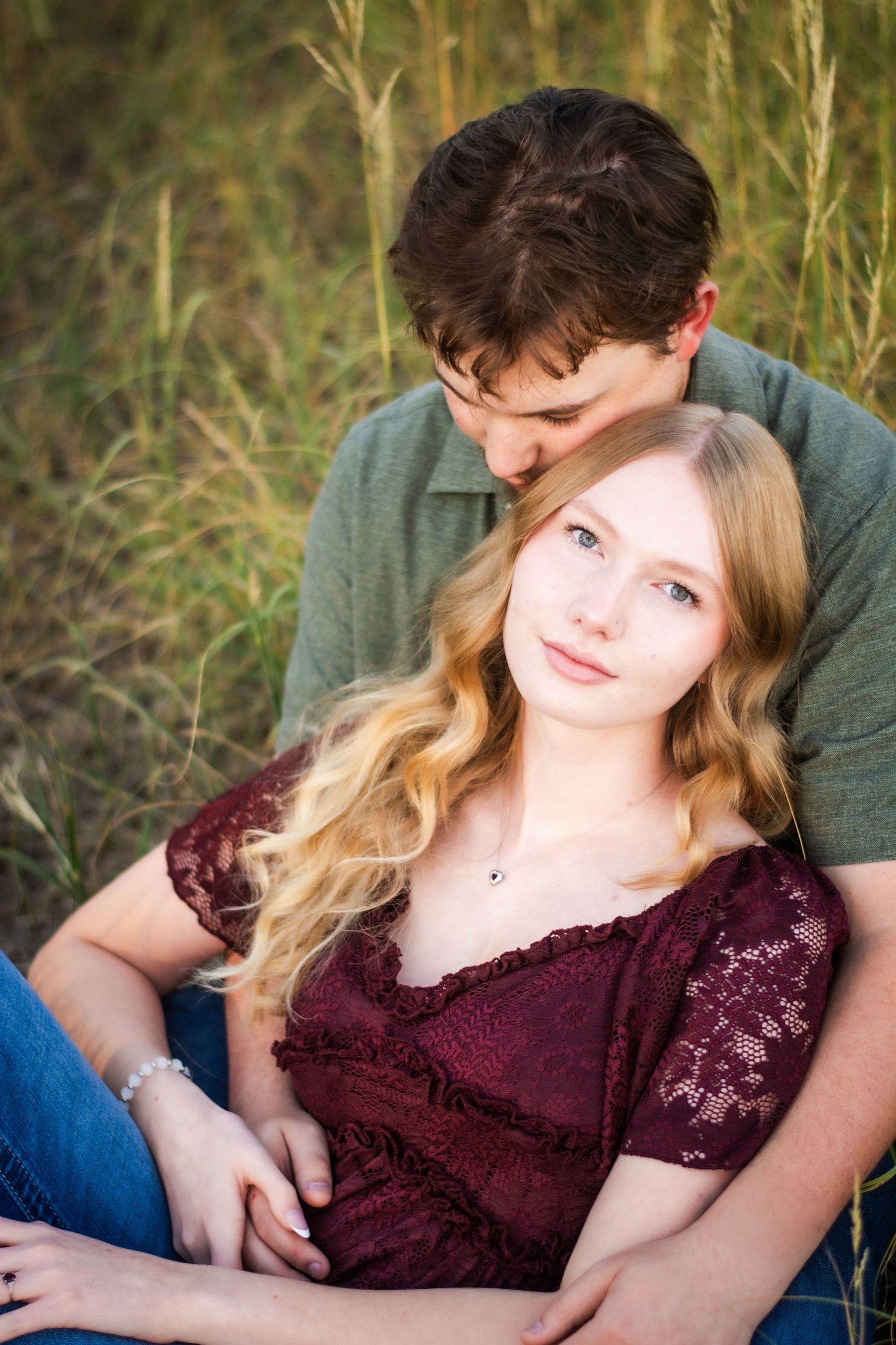 Young blonde woman in burgundy lace top leans against man in green shirt in tall grass.