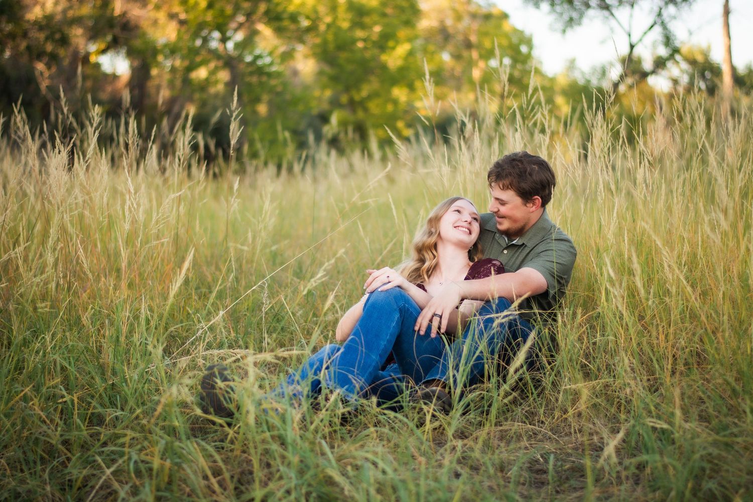 Happy couple sits together laughing in a golden field of tall grass during engagement session.
